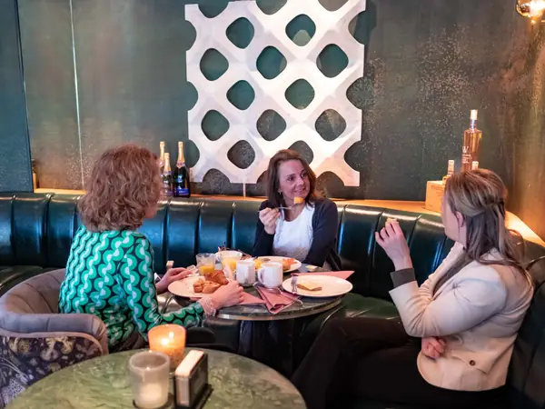Drie vrouwen ontbijten op groene bank, lachend gesprek