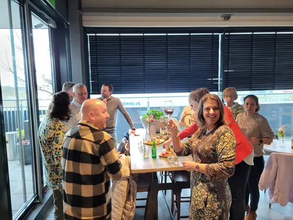 Lachende vrouw in pauwenjurk heft wijnglas, groep aan statafel
